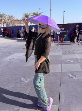 The image features a woman standing outside, wearing sunglasses and holding an umbrella over her head. She appears to be posing for a photo or video. The background of the image shows several people walking around, possibly attending an event or gathering. There is also a sign in the scene that reads "Massive congratulations to Arcium on their launch! Arcium has always stood out to me as one of the most innovative & giga brained technologies on SOLANA".