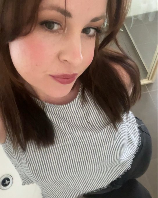 A selfie of a woman with long brown hair, wearing a striped sleeveless top, looking directly at the camera.