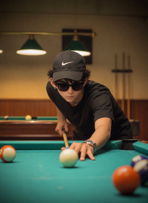 A person wearing a black Nike cap, sunglasses, and black shirt playing pool, aiming at the cue ball on a green table with billiard balls scattered around.