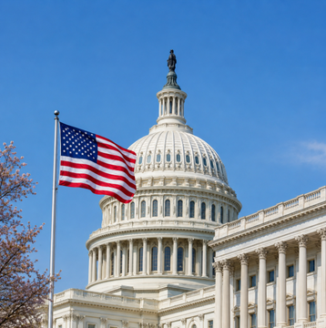 The image shows the U.S. Capitol dome with an American flag waving in the foreground against a clear blue sky, symbolizing the legislative backdrop to the political negotiations over the CLARITY Act described in the post. The image visually anchors the story about Democratic and Republican lawmakers debating the bill's ethics provisions in the halls of Congress.