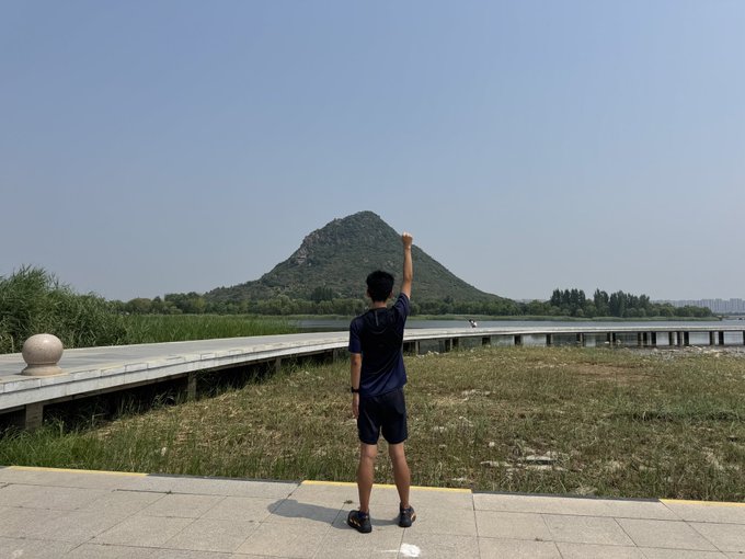 A man stands on a paved path with his fist raised triumphantly toward a mountain in the distance, appearing to pose after a run. The scenic outdoor setting with a boardwalk and lake aligns with the post about running with his son, likely captured during a STEPN move-to-earn activity.