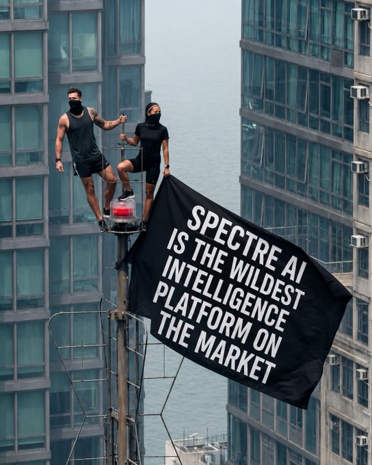 Two masked individuals stand atop a pole between skyscrapers holding a black banner reading 'SPECTRE AI IS THE WILDEST INTELLIGENCE PLATFORM ON THE MARKET'.
