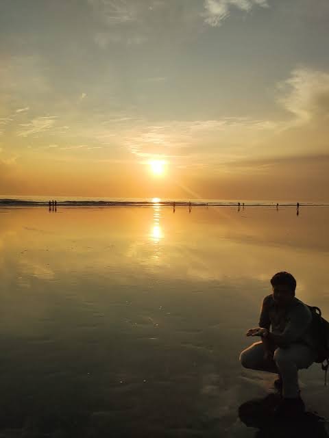 A silhouetted figure crouches on a wet, mirror-like beach at sunset while other small figures stand in the shallow water reflecting the golden sun.
