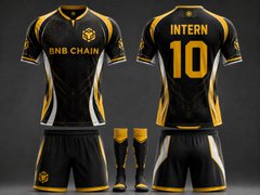 The image features a black and gold soccer uniform, likely representing a professional soccer team or a sponsor. The uniform has yellow accents that make it stand out against the dark background. In the foreground of the image, there are two pairs of shorts - one pair is positioned on top of the other. These shorts also have yellow accents, adding to the overall color scheme of the outfit. The text "BNB Chain" and "Intern 10" can be seen in the image, possibly indicating the team's name or their number.