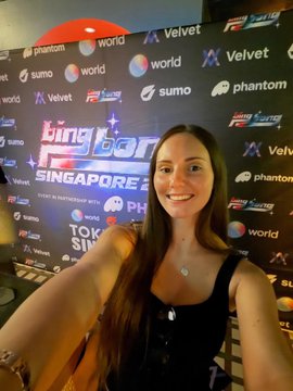 A selfie of a smiling young woman with long dark hair wearing a black top and a necklace, taken in front of a step-and-repeat backdrop displaying logos like 'phantom', 'velvet', 'sumo', and 'Bingo Bonanza Singapore'.