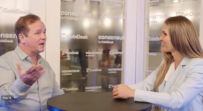 Two people are seated at a small round table during an interview at a CoinDesk 'Consensus' event.