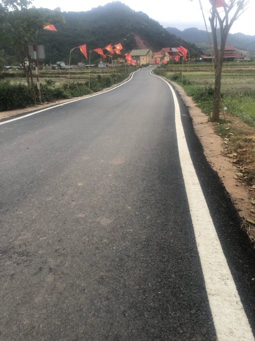 A newly paved road winds through a rural Vietnamese landscape, lined with red flags and leading toward a small village with traditional houses nestled against misty mountains. The image complements the post's message about building a stronger Vietnamese community, symbolizing progress and a fresh start on the journey ahead.