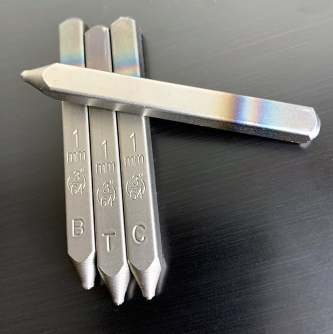 Three metal Bitcoin stamp tools labeled "B," "T," and "C" with "1 mm" and "3/64" markings lie crossed on a dark surface, illustrating the post's message about personally stamping and verifying one's own seed phrase recovery process rather than relying on third-party solutions.