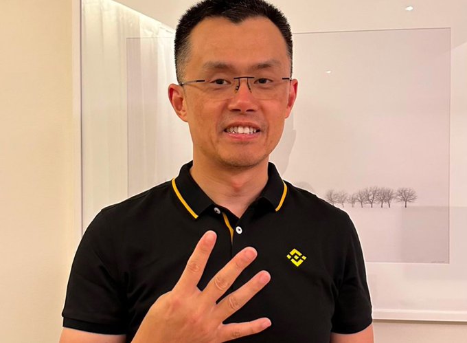 A man wearing glasses and a black polo shirt with the Binance logo smiles and holds up three fingers, appearing to indicate the number 3 in reference to the low count of Cardano nodes producing blocks on the mainnet.