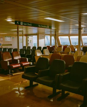 An empty ferry interior with rows of dark red and black seats, featuring illuminated directional signs reading "Sun Deck" and "No Smoking" hanging from the ceiling.