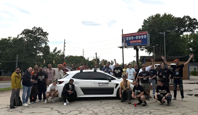 A group of around 20 people pose around a white car displaying a "SPX6900" license plate, with a billboard in the background advertising "Car, Truck, SUV, CYCLE INJURY 999-9999 George Sink Injury Lawyers." The image captures a community gathering celebrating the #SPX6900 SC 2026 event, with attendees showing enthusiasm as they surround the branded vehicle.
