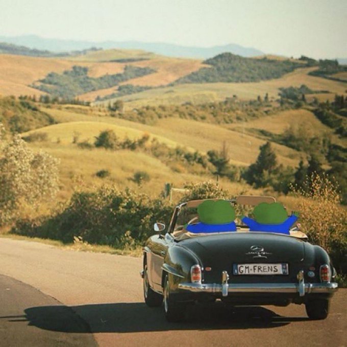 A dark green Mercedes-Benz convertible with two cartoon-sticker passengers cruises down a winding road through Tuscan-style rolling hills, its license plate reading "GM·FRENS." The carefree road-trip imagery visually echoes the post's optimistic "just another week to go harder" sentiment, while the playful stickers reflect the casual, community-driven "woke up on-chain" crypto vibe.