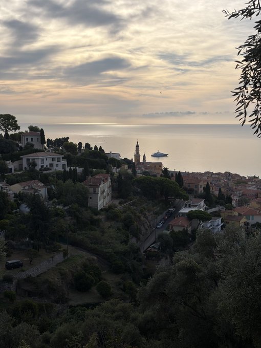 An elevated scenic view of a coastal Mediterranean town at sunset, featuring terracotta-roofed buildings, a church tower, lush greenery, and a large cruise ship on the calm sea in the distance.
