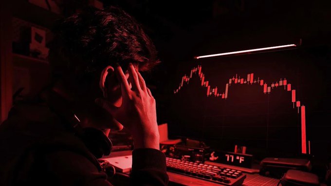 A man in a dark room bathed in red light holds his head in his hand while looking at a computer monitor displaying a downward-trending red candlestick financial chart.