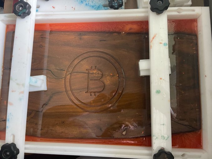 An overhead view of a wooden tray filled with water and orange resin, featuring a Bitcoin logo etched into the center.