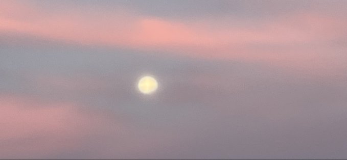 A bright moon or sun glowing through a soft sky painted with pink and blue clouds at twilight.