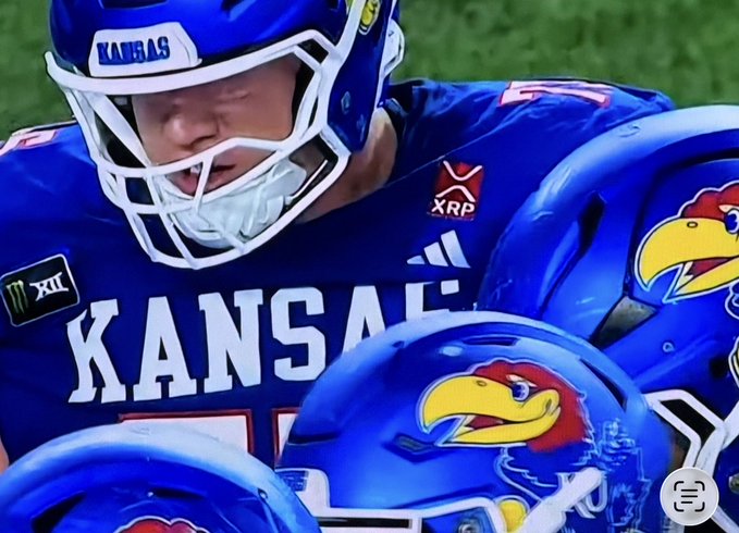 A close-up of Kansas football players in blue uniforms with the Jayhawk logo, with XRP branding visible.