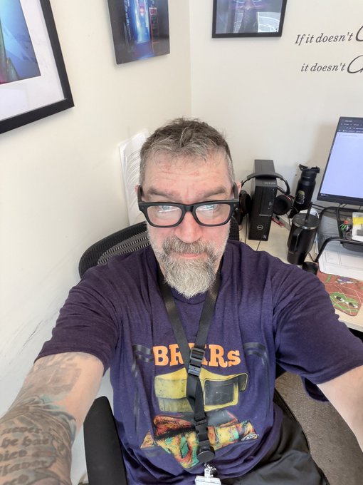 A bearded man with glasses and a tattooed arm wearing a purple graphic t-shirt, sitting at a desk with computer equipment behind him.