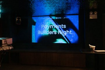 A screen displays the text "Payments Builders Night" at an event venue, with speakers mounted on the wall and a stage visible below.
