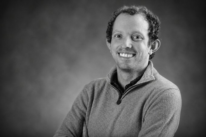 The image features a man wearing a gray sweater, smiling warmly as he poses for the camera. The photo has been edited into black and white, giving it an artistic touch. The background of the image is a dark color, which contrasts with the man's attire and adds depth to the photograph. The caption below the image reads: "Really sad to hear about the passing of Nathan Allman, founder of @OndoFinance. He played a major role in pushing the RWA sector forward and helping bridge traditional finance with on-chain markets."