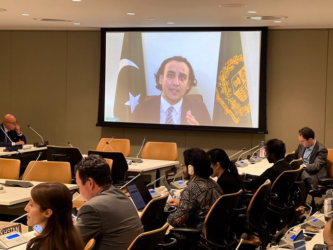 A conference room scene with attendees seated around a table while a large screen displays a man in a suit, with the Pakistan flag visible behind him and another emblem beside him.