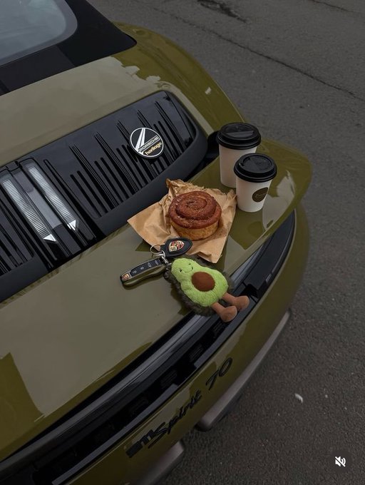 Two takeaway coffee cups, a cinnamon roll on parchment paper, keys with an avocado plush keychain, and a Volkswagen logo arranged on the hood of an olive-green Volkswagen Scirocco 1.8.