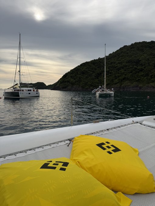 Two yellow life jackets sit on the deck of a boat overlooking a calm bay with two sailboats anchored near a green, hilly coastline under a cloudy sky.