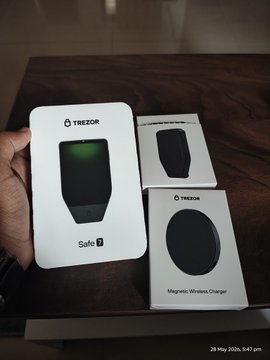 The image shows a person holding two boxes of a product called "@Trezor Safe" along with a white box containing a black device. The boxes are placed on a wooden table, and there's also an electronic device visible in the background. The text in the image suggests that these items were recently unboxed or received as gifts, possibly for use with a smartphone.