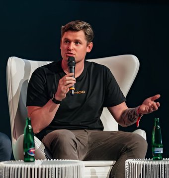 A man in a black 'ROC' branded t-shirt speaking into a microphone while seated on a white chair at what appears to be a conference or panel discussion.