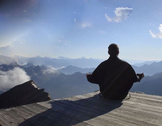 A silhouetted figure sitting in a meditative pose on a wooden deck overlooking a vast mountain range with clouds below.