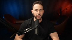 A bearded man with dark hair wearing a black t-shirt, sitting in front of a microphone in a studio with red and blue lighting.