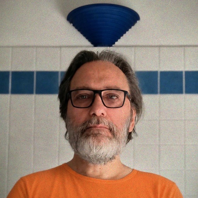 A close-up portrait of a middle-aged man with glasses, a gray beard, and an orange shirt, standing in a bathroom with white and blue tiled walls beneath a blue ceiling lamp.