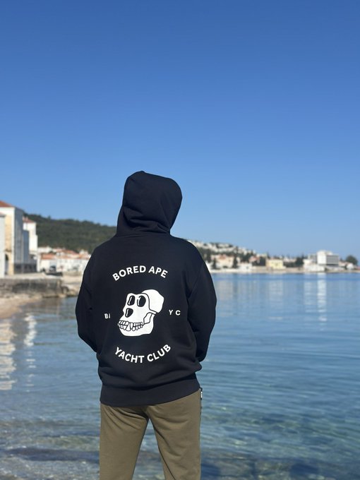 The image features a person standing near water, wearing a black hoodie with white text that reads "GM Bored Ape Yacht Club" and "Yacht Club". The person appears to be looking out at the ocean or sea, possibly enjoying their surroundings.