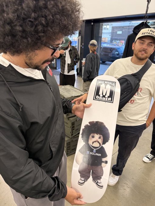 A man with an afro holds a white skateboard deck featuring a cartoon character design with "NEW WORLD M ORDER" and "SOLRAW" text, while another man wearing an "I AM RETARDED" cap stands beside him in what appears to be a skate shop. The image ties to the testing teaser for "$Miu," showcasing the branded skateboard merch collaboration.