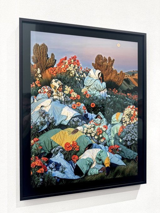 A framed art print hanging on a white wall depicts a vibrant, surreal scene of figures in colorful hooded garments reclining among lush flowers and cacti, with rolling hills and a moon in the background.