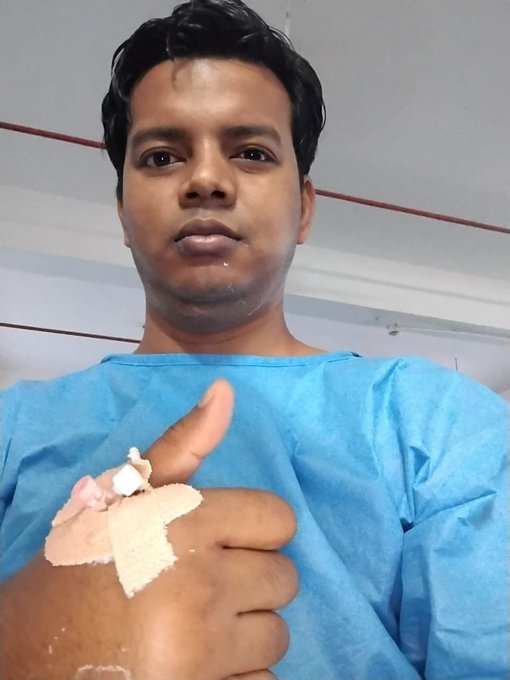 A young man in a blue hospital gown gives a thumbs-up while showing bandaged and stitched injuries on his hand.