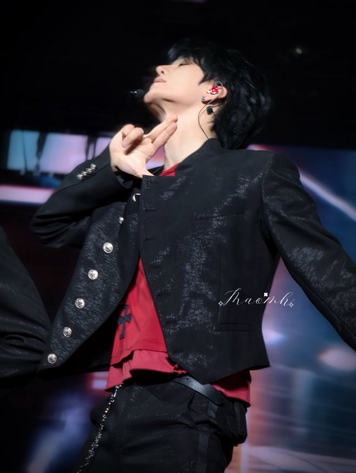 A performer on a dimly lit stage tilts their head back with eyes closed, wearing a black patterned jacket over a red shirt, a red in-ear monitor, and a small black headset microphone, with a watermark reading 'Haonhi'.