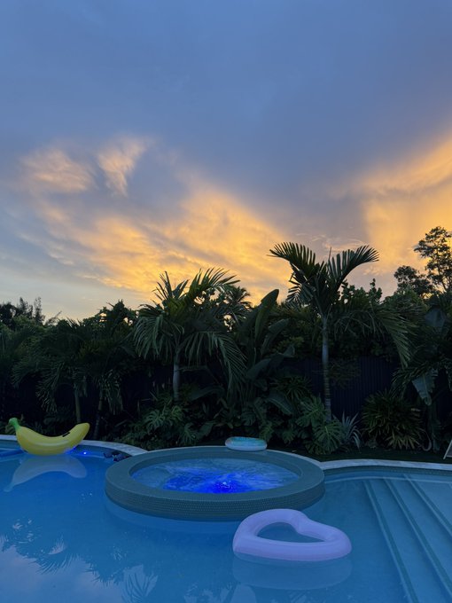 A serene sunset scene over a backyard swimming pool featuring an integrated jacuzzi with blue lighting, surrounded by tropical palm trees, with a yellow banana-shaped float and a pink heart-shaped float resting on the water.