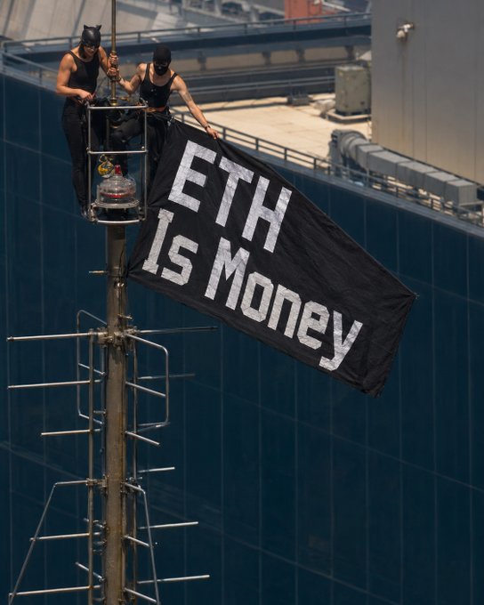 Two protesters wearing black masks and tank tops hang a large black flag reading "ETH IS MONEY" from the antenna of a high-rise building, with a glass skyscraper visible in the background.