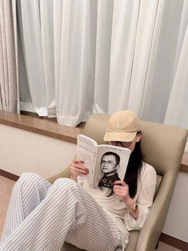 A person wearing a cap and striped pants sits in a chair reading a book featuring a black-and-white portrait of a man on the cover.