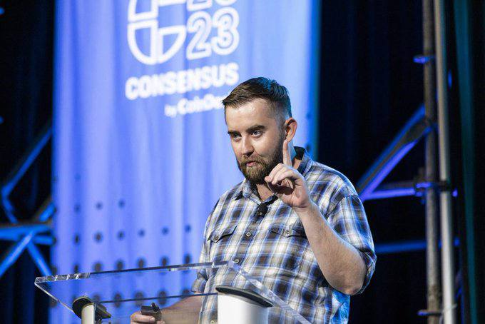 A man with a beard in a plaid shirt speaking at a podium during the 'CONSENSUS by CoinDesk 23' event, gesturing with his hand.