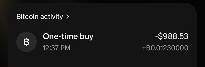 A dark-themed mobile app screen displays a Bitcoin activity log showing a one-time buy of -$988.53 for ₿0.01230000 at 12:37 PM.
