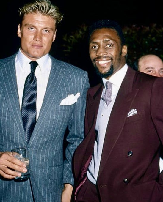 Two men in suits—one in a blue pinstripe suit holding a glass, and the other in a maroon double-breasted suit—smiling together at what appears to be a formal event.