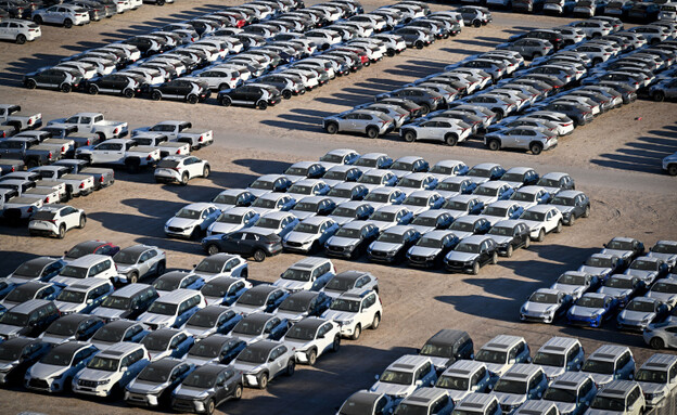 An aerial view of a large parking lot or storage yard densely packed with rows of newly manufactured cars, mostly white and silver sedans and SUVs.