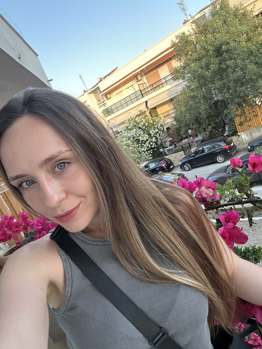 A selfie of a young woman with long brown hair wearing a gray tank top, taken outdoors with a building, parked cars, and pink flowers in the background.
