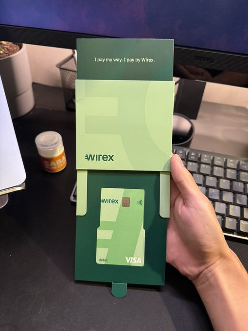 A hand holds a green Wirex debit card package displaying the VISA debit card, with the tagline 'I pay my way. I pay by Wirex' on the desk next to a keyboard and monitor.