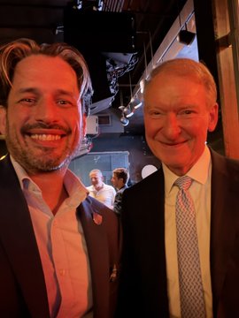 A photo of two men smiling together at what appears to be an indoor event, one wearing a dark suit and the other in a dark blazer with a patterned tie.