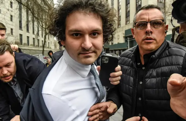 A man with curly hair in a suit and tie is escorted by another man in a black puffer jacket who is holding a smartphone, appearing to be in a crowded outdoor urban setting.