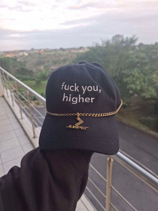A navy blue baseball cap with the embroidered text 'fuck you, higher' and a gold chain attached is displayed against a scenic outdoor backdrop.