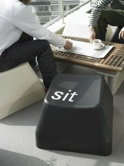 Two people sit at a low outdoor table having coffee, while a black cube-shaped stool in the foreground has the word 'sit' written on it in white letters.